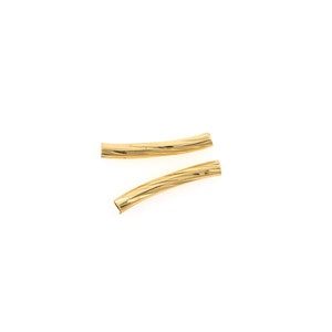 Puede incluir: Dos cuentas tubulares doradas y curvadas con una superficie texturizada y acanalada. Las cuentas están colocadas en diagonal sobre un fondo blanco. Estos componentes de joyería son adecuados para crear collares, pulseras o pendientes.