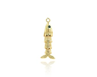 Könnte beinhalten: Goldfarbener Fischanhänger mit einem kleinen grünen Edelstein als Auge. Der Anhänger hat ein detailliertes, geschichtetes Design und eine Schlaufe oben für eine Kette. Die Fischschwanzflosse ist unten sichtbar.