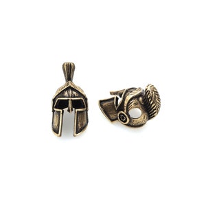 May include: Two gold-toned metal Spartan helmet charms. One charm is a full helmet with a plume on top, the other is a side profile of a helmet.
