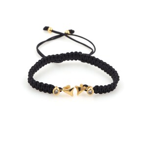 Pode incluir: Um bracelete trançado preto com pingentes de flecha dourados e detalhes de cristal transparente.