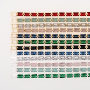 May include: Rows of colorful rectangular gemstones in various colors, including red, green, blue, and clear, set in gold and silver metal settings. The gemstones are arranged in a grid pattern against a white background, creating a vibrant and eye-catching design.