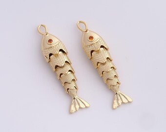 Könnte beinhalten: Zwei goldfarbene Anhänger in Fischform. Jeder Anhänger hat einen strukturierten Körper, einen kleinen orangefarbenen Edelstein für das Auge und eine Schlaufe zum Aufhängen. Die Fische haben ein detailliertes Flossendesign.