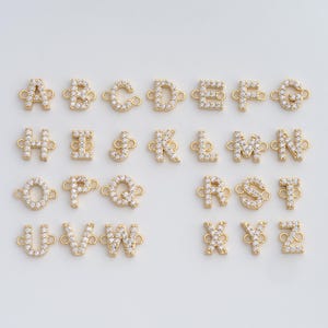 May include: A collection of gold-colored alphabet charms, each encrusted with small, clear crystals. The letters A through Z are displayed, each with loops for attachment. The charms are arranged in neat rows against a plain background.