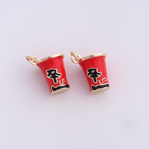 May include: Two red enamel charms shaped like ramen cups with gold trim. Each charm features a black graphic and a gold loop for attaching to jewelry. The charms are set against a white background.