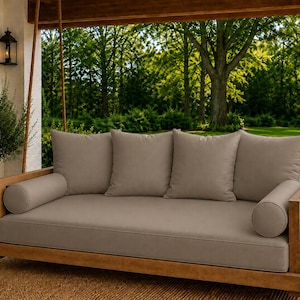 May include: A wooden porch swing bed featuring a natural light brown wood frame suspended by thick ropes. It includes a large rectangular seat cushion, four square back cushions, and two cylindrical bolster pillows, all in a solid beige color.