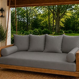 May include: A wooden porch swing bed featuring a natural light brown wood frame suspended by thick ropes. The swing includes a large rectangular seat cushion, four square back cushions, and two cylindrical bolster pillows, all in a solid gray.