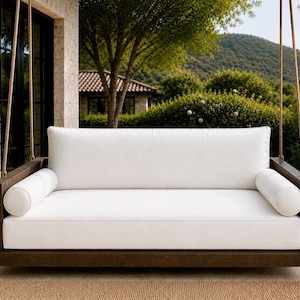 May include: A hanging porch swing featuring a dark brown wooden frame and thick white cushions. The rectangular seat includes a long back cushion and two cylindrical bolster pillows on each side, suspended by thick ropes from an overhead structure.