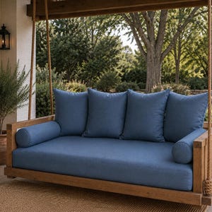 May include: A wooden porch swing featuring a natural brown finish, suspended by thick ropes. The swing includes a rectangular seat cushion, four square back cushions, and two cylindrical bolster pillows, all upholstered in a solid blue fabric.
