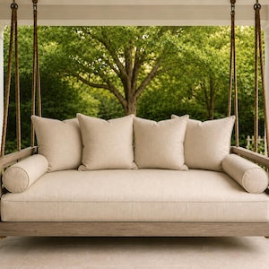May include: A rectangular wooden porch swing bed suspended by thick ropes, featuring a light beige mattress, two cylindrical bolster pillows at the ends, and four square beige throw pillows arranged across the back of the seat frame.