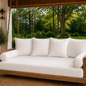 May include: A wooden hanging daybed suspended by thick ropes, featuring a rectangular frame with a natural wood finish. The daybed includes a thick seat cushion, four square back cushions, and two cylindrical bolster pillows, all in white.