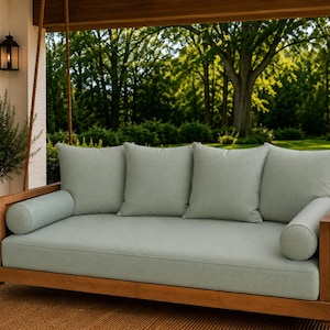 May include: A wooden porch swing featuring a rectangular frame suspended by thick ropes. The swing includes a large seat cushion, three square back cushions, and two cylindrical bolster pillows, all upholstered in a solid light sage green fabric.