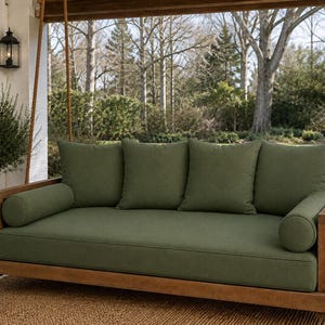 May include: A wooden porch swing featuring a rectangular frame suspended by thick ropes. The swing includes a large seat cushion, three square back cushions, and two cylindrical bolster pillows, all upholstered in a solid olive green fabric.