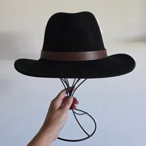 Pu&ograve; includere: Un cappello fedora nero caratterizzato da una fascia in pelle marrone liscia che circonda la base della corona. Il cappello presenta una corona strutturata e pizzicata e una tesa larga e piatta, offrendo una silhouette classica e uniforme.