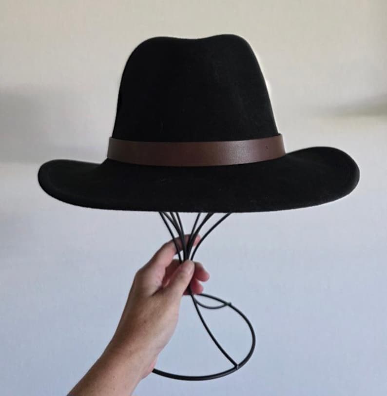 Pu&ograve; includere: Un cappello fedora nero caratterizzato da una fascia in pelle marrone liscia che circonda la base della corona. Il cappello presenta una corona strutturata e pizzicata e una tesa larga e piatta, offrendo una silhouette classica e uniforme.