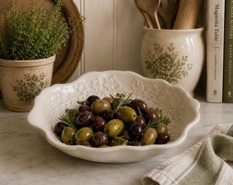 May include: A cream-colored ceramic bowl with a scalloped edge and embossed floral detailing, filled with a mixture of green and dark purple olives garnished with fresh green rosemary sprigs, presented on a white marble surface.