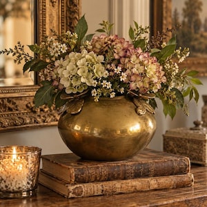 May include: A round, polished brass vase featuring decorative leaf accents at the rim, filled with a lush arrangement of white and mauve hydrangea blossoms, small white flowers, and green foliage, creating a dense, rounded floral display.