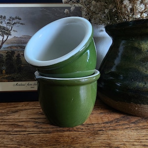 Może przedstawiać: Dwie małe, zielone ceramiczne miski z białym wnętrzem ułożone na drewnianej powierzchni. Miski mają błyszczące wykończenie i zaokrąglony kształt. W tle ciemnozielona waza oraz oprawiony krajobraz.