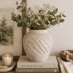 May include: A round, textured beige ceramic vase with diagonal embossed stripes, filled with a bouquet of green eucalyptus leaves and small white baby's breath flowers, resting on a stack of two books titled Live Beautifully and This Is Home.