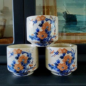 May include: Three ceramic cups with a white speckled finish. Each cup is decorated with orange and blue floral patterns. The cups are stacked on top of each other, showcasing the design and shape of the cups.
