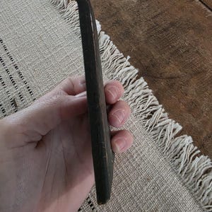 Puede incluir: Un objeto de madera alargado de color marr&oacute;n oscuro, posiblemente una herramienta o una pieza decorativa. El objeto se sostiene en una mano, centr&aacute;ndose en su forma y color. La madera parece envejecida, con una textura ligeramente desgastada.