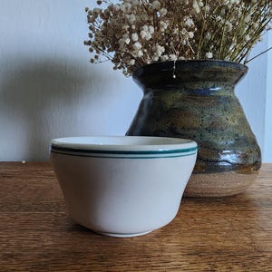 May include: A small, white ceramic bowl with a green stripe near the rim. The bowl sits on a wooden surface, with a decorative vase and dried flowers in the background. The vase is dark with a textured, mottled appearance.