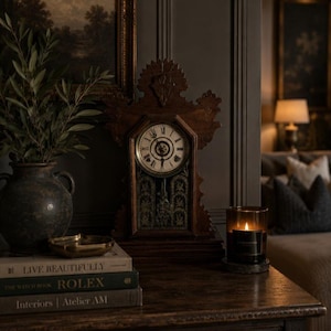 May include: A dark wooden mantel clock with a circular face and ornate carved top, positioned next to a stack of three books titled Live Beautifully, The Watch Book Rolex, and Interiors Atelier AM, a dark ceramic vase, and a lit candle.