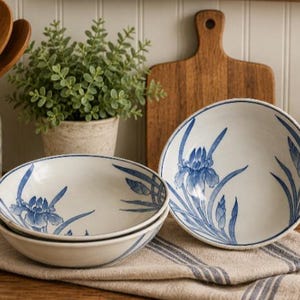 May include: Three ceramic bowls featuring a hand-painted blue iris floral pattern on a white background. Two bowls are stacked together, while one bowl stands upright to the right, all resting on a folded beige cloth with thin blue stripes.