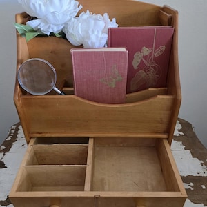 May include: A wooden desk organizer with a drawer and multiple compartments. It holds two red books, a magnifying glass, and white flowers. The organizer is made of light-colored wood and has a curved top.