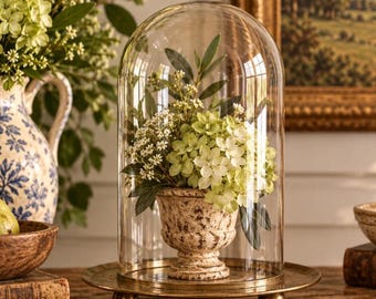 Può includere: Una cupola in vetro trasparente poggia su un vassoio rotondo in ottone con piccoli piedini a sfera. All'interno, un'urna beige dall'aspetto in pietra contiene un bouquet di fiori di ortensia verdi e foglie scure.