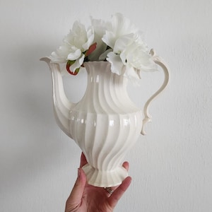Puede incluir: Un jarrón en forma de jarra de color crema, de estilo vintage, con un diseño en espiral, que contiene flores blancas. El jarrón tiene un asa y una base decorativa. Las flores son blancas con acentos verdes y rojos.