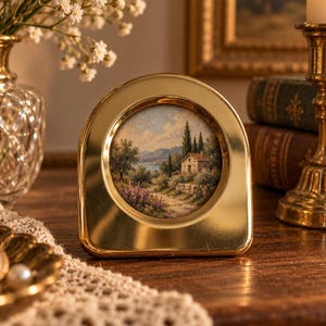 May include: A polished gold-toned metal picture frame with an arched top, containing a circular landscape painting of a stone cottage, cypress trees, and rolling hills under a blue sky with soft white clouds.
