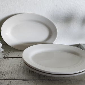 May include: Two white oval serving platters and a stack of two round plates are displayed on a distressed, white-painted wooden surface. The platters are in the background, and the plates are in the foreground.
