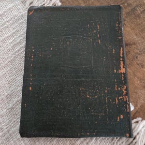 Puede incluir: Un libro rectangular verde oscuro, encuadernado en cuero, con un aspecto desgastado. La cubierta muestra un dise&ntilde;o en relieve y zonas de desgaste, revelando un tono m&aacute;s claro debajo. El libro est&aacute; colocado sobre una superficie texturizada blanquecina.
