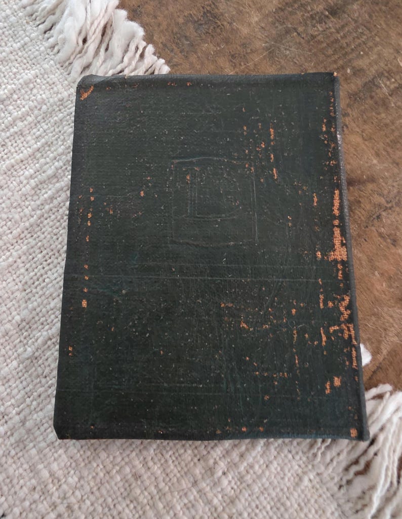 Puede incluir: Un libro rectangular verde oscuro, encuadernado en cuero, con un aspecto desgastado. La cubierta muestra un dise&ntilde;o en relieve y zonas de desgaste, revelando un tono m&aacute;s claro debajo. El libro est&aacute; colocado sobre una superficie texturizada blanquecina.