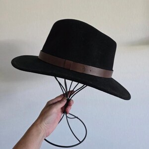 Pu&ograve; includere: Un cappello fedora nero caratterizzato da una tesa larga e una fascia in pelle marrone liscia avvolta attorno alla base della corona. Il cappello presenta una struttura con parte superiore pizzicata e tesa piatta, dal design pulito.