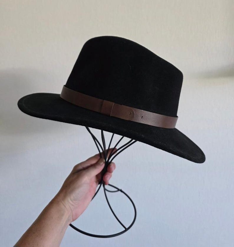 Pu&ograve; includere: Un cappello fedora nero caratterizzato da una tesa larga e una fascia in pelle marrone liscia avvolta attorno alla base della corona. Il cappello presenta una struttura con parte superiore pizzicata e tesa piatta, dal design pulito.