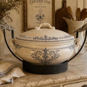 Pode incluir: Uma terrina de cerâmica cor de creme com tampa, apresentando padrões florais azuis e duas pegas laterais. Repousa num suporte de metal preto com detalhes prateados nas laterais, proporcionando uma base estável para o recipiente de servir.