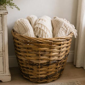 Puede incluir: Una cesta grande, redonda y tejida de mimbre marrón llena de varias mantas de punto de color crema dobladas. Las mantas presentan patrones texturizados y detalles de flecos, creando una apariencia suave y superpuesta dentro de la cesta.