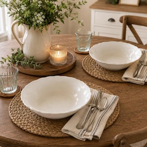 May include: A wooden dining table set for two with white ceramic bowls, silver forks and spoons, and linen napkins with gray stripes. A white pitcher filled with greenery, a glass candle holder, and two clear drinking glasses are present.