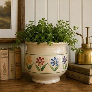 May include: A cream-coloured ceramic planter with a rounded body and a flared rim, featuring a hand-painted horizontal green line and a repeating pattern of yellow, red, blue, and purple flowers with green leaves around the centre.