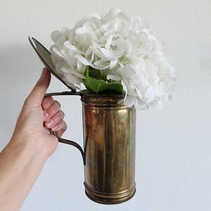 Puede incluir: Un jarrón de latón vintage con asa, que contiene un ramo de hortensias blancas. El jarrón tiene forma cilíndrica con estrías verticales y una tapa con bisagras. Las flores son abundantes, contrastando con el latón envejecido.