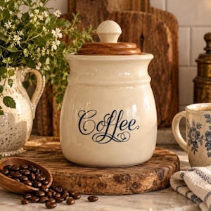May include: A cream-colored ceramic canister with a rounded body and a dark wooden lid featuring a knob handle. The word Coffee is printed on the front in a dark blue cursive script. The canister has a smooth finish and a wide opening.