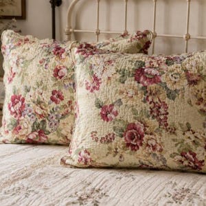May include: Two square quilted pillow shams featuring a cream-colored base with a dense floral pattern in shades of deep red, muted pink, yellow, and green. The fabric has a visible stitched texture throughout, and the edges are finished with a simple hem.