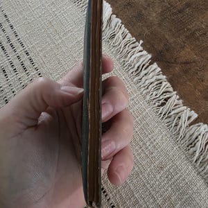 Puede incluir: Un libro peque&ntilde;o, rectangular y antiguo con una cubierta verde oscuro y p&aacute;ginas amarillentas. El libro se sostiene en una mano, mostrando su grosor y desgaste. El fondo es una superficie texturizada de tonos neutros.