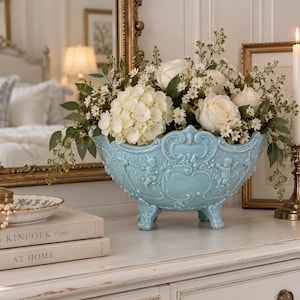 Puede incluir: Un cuenco de cerámica azul claro con relieves ornamentales de querubines y volutas, sostenido por cuatro patas pequeñas. El cuenco contiene un arreglo floral de rosas blancas, hortensias blancas y follaje verde.