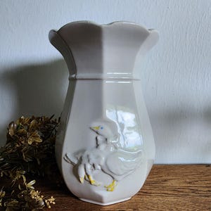 May include: A cream-colored ceramic vase with a scalloped rim and a flared, faceted body. The vase features a raised relief design of two geese with yellow feet and blue eyes. The vase is displayed on a wooden surface.