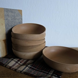 Pode incluir: Seis pequenas tigelas de madeira castanha clara, uma separada. As tigelas ovais têm um acabamento liso e natural. Um livro antigo está encostado na pilha. Um pano xadrez está sob as tigelas. Adequado para lanches.