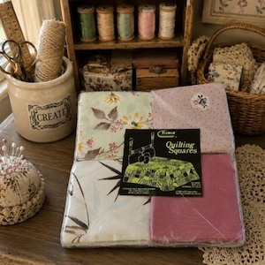 May include: A pack of Remar Quilting Squares containing assorted fabric patterns, including floral and solid pink, with a label showing a quilt illustration. The set is displayed alongside a ceramic jar with a ruler, scissors, and twine.