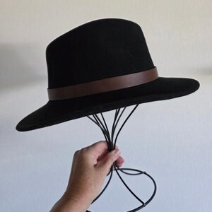 Pu&ograve; includere: Un cappello fedora nero caratterizzato da una fascia liscia marrone scuro che circonda la base della corona. Il cappello presenta una tesa strutturata e una parte superiore arrotondata, esposto su un supporto in filo metallico nero.