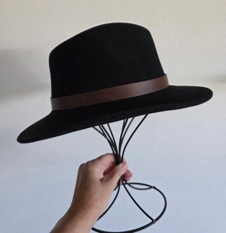 Pu&ograve; includere: Un cappello fedora nero caratterizzato da una fascia liscia marrone scuro che circonda la base della corona. Il cappello presenta una tesa strutturata e una parte superiore arrotondata, esposto su un supporto in filo metallico nero.